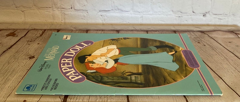 May include: A vintage Disney's The Little Mermaid paper doll book with a colourful cover featuring Ariel and Prince Eric. The cover has the text "Paper Doll" in a purple and white circle and "Disney's The Little Mermaid" in a blue and white rectangle.