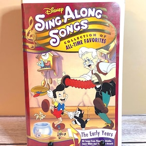 May include: A Disney Sing Along Songs VHS tape, featuring Pinocchio and other characters. The cover is red and white with the title "Sing Along Songs" and "Collection of All-Time Favorites". The Early Years is printed on the bottom.