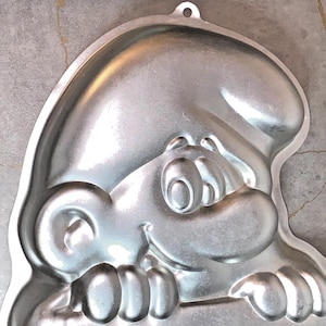 May include: A silver-colored Smurf-shaped cake pan. The pan features a detailed embossed design of a Smurf's face, including eyes, nose, and a smiling mouth. The pan has a loop at the top for hanging and is made of a shiny metal.