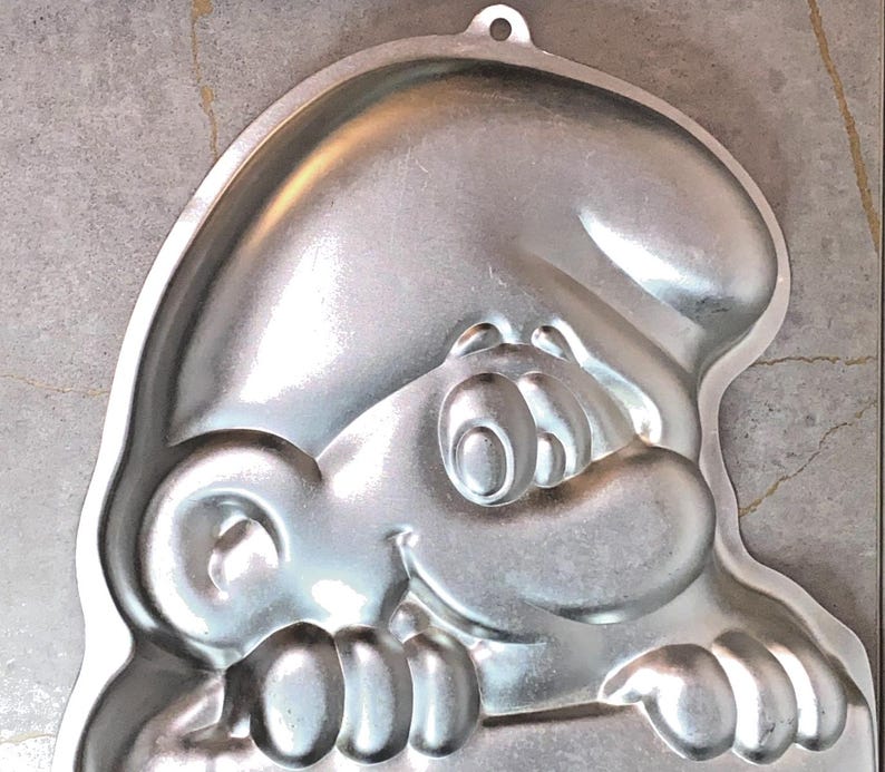 May include: A silver-colored Smurf-shaped cake pan. The pan features a detailed embossed design of a Smurf's face, including eyes, nose, and a smiling mouth. The pan has a loop at the top for hanging and is made of a shiny metal.