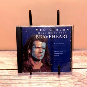 May include: A CD case for the movie "Braveheart" featuring Mel Gibson. The cover has a blue background with a portrait of Gibson with blue face paint. The text "Mel Gibson More Music From Braveheart" is displayed.