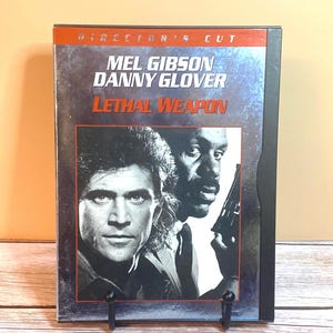 May include: DVD case for the movie "Lethal Weapon" featuring Mel Gibson and Danny Glover. The cover has a gray and black background with the movie title in red. The case is displayed on a black stand.