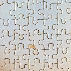 以下が含まれることがあります： 部分的に組み立てられたジグソーパズルのクローズアップ。パズルのピースはオフホワイトで、小さなシミがいくつかあります。ピースはクラシックな形状で組み合わされ、グリッドのようなパターンを作り出しています。画像はパズルのテクスチャとデザインを強調しています。
