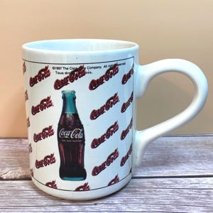 Puede incluir: Una taza de cerámica blanca con un patrón repetitivo del logotipo rojo de Coca-Cola y una ilustración central de una botella de vidrio clásica. El texto en la parte superior dice 1997 The Coca-Cola Company. All rights reserved. Tous droits réservés.