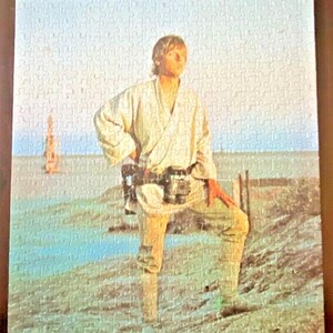 以下が含まれることがあります： スターウォーズのシーンを特徴とする完成したジグソーパズル。画像には、ベージュの衣装を着た人物が砂地の表面に立っており、青い空と背景に構造物があります。パズルは多くの小さなピースで構成されています。
