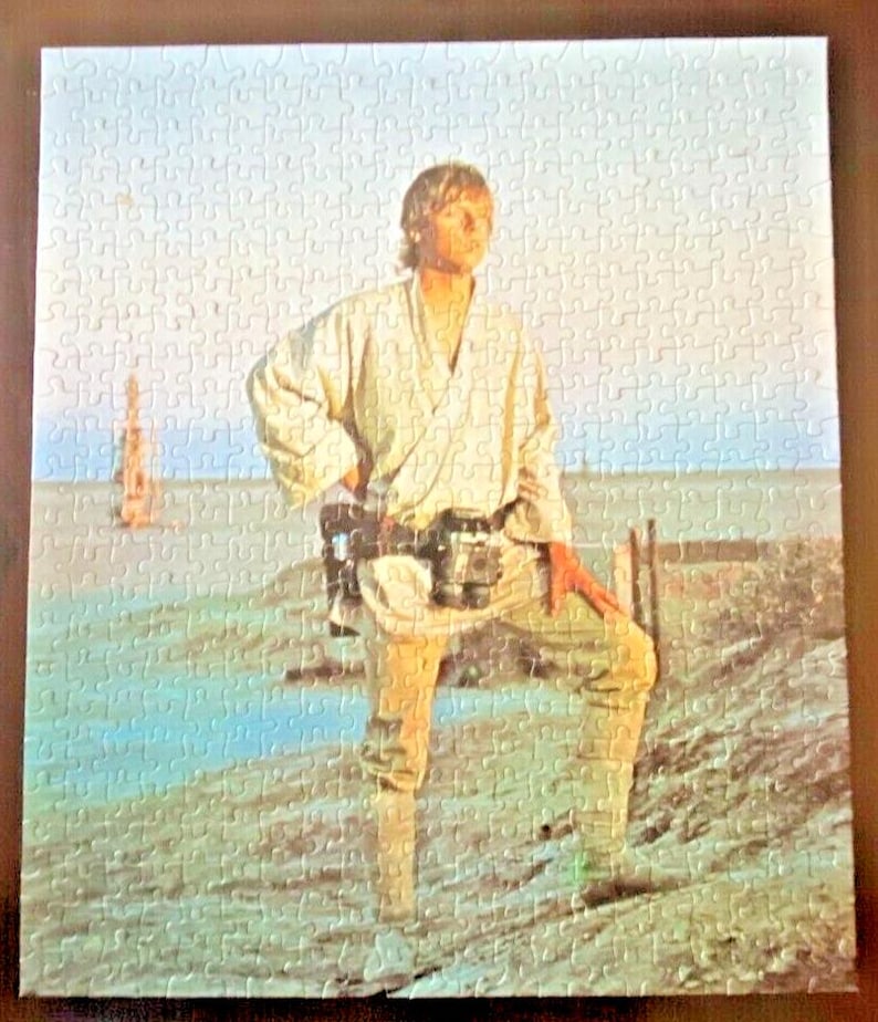 以下が含まれることがあります： スターウォーズのシーンを特徴とする完成したジグソーパズル。画像には、ベージュの衣装を着た人物が砂地の表面に立っており、青い空と背景に構造物があります。パズルは多くの小さなピースで構成されています。