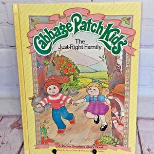 May include: A yellow book with a green and pink Cabbage Patch Kids logo. The title is "The Just-Right Family". The cover features an illustration of two Cabbage Patch Kids dolls walking in a forest setting. The book is a Parker Brothers Story Book.