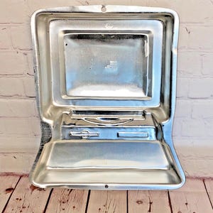 May include: A silver metal cake pan shaped like a vintage computer. The pan has a rectangular screen, a slot for a floppy disk, and a small handle.