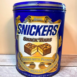 May include: A cylindrical blue metal tin featuring the Snickers logo, the text Snack Bars, Packed With Peanuts, and Snickers Really Satisfies. The front displays an illustration of two halved chocolate bars. It states 2 One Lb. Bags (Net Wt 32 Oz.).