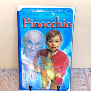 Puede incluir: Una cinta VHS azul de "Las Aventuras de Pinocho" con Martin Landau y Jonathan Taylor Thomas. La portada tiene una imagen vibrante de los personajes y el título en rojo. La cinta está sostenida por un soporte de metal negro.