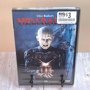May include: Sealed DVD of the horror film "Hellraiser" by Clive Barker. The cover features a character with pins in their head, with the title in red letters. The DVD is in a plastic case and has a price sticker.