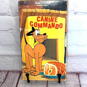 May include: A vintage VHS tape of "Canine Commando" featuring Pluto from Walt Disney Home Video. The cover has a yellow background with Pluto and a ham. The tape is displayed on a black metal stand against a white and wood background.