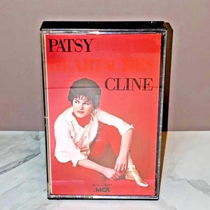May include: A vintage cassette tape featuring the album "Patsy Cline Heartaches." The cover is predominantly red, with the artist's name and album title in white and orange. A black and white photo of Patsy Cline is featured on the cover.