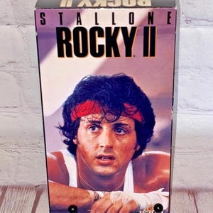 Puede incluir: Una caja de cinta VHS para la película "Rocky II" protagonizada por Sylvester Stallone. La caja presenta una imagen de primer plano de Stallone como Rocky Balboa, con un pañuelo rojo en la cabeza y un tanque blanco. El texto "Stallone" y "Rocky II" está impreso en negro sobre un fondo blanco.