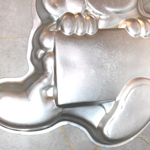 May include: A silver-colored, bear-shaped cake pan. The pan is made of metal and features a rectangular area held by the bear's paws. The bear's body and legs are visible, with rounded features and a smooth surface. The pan is designed for baking cakes.