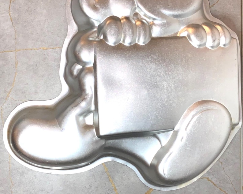 May include: A silver-colored, bear-shaped cake pan. The pan is made of metal and features a rectangular area held by the bear's paws. The bear's body and legs are visible, with rounded features and a smooth surface. The pan is designed for baking cakes.