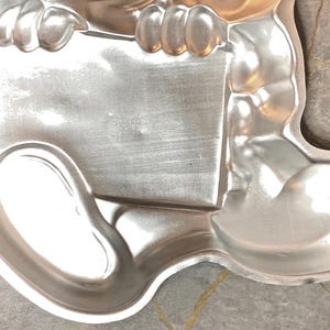 May include: A silver-colored, metal baking pan in the shape of a cartoon character. The pan has a rectangular section held by two rounded arms, and a rounded foot section. The pan is made of a shiny metal and is designed for baking.
