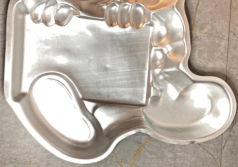 May include: A silver-colored, metal baking pan in the shape of a cartoon character. The pan has a rectangular section held by two rounded arms, and a rounded foot section. The pan is made of a shiny metal and is designed for baking.
