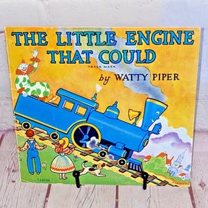 May include: Vintage children's book cover featuring a blue train engine with a smiling face. The cover is yellow with the title "The Little Engine That Could" in red and black lettering. The book is by Watty Piper.