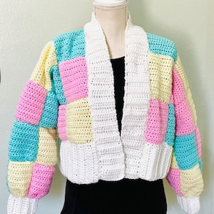May include: A white crocheted cardigan with a patchwork design in pink, yellow, and blue. The cardigan has a long, open front and long sleeves.