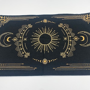 Puede incluir: Tela rectangular negra con un dise&ntilde;o celestial dorado. El dise&ntilde;o incluye un sol central, lunas crecientes, estrellas y formas geom&eacute;tricas. La tela probablemente se utiliza para el tarot u otras pr&aacute;cticas espirituales.