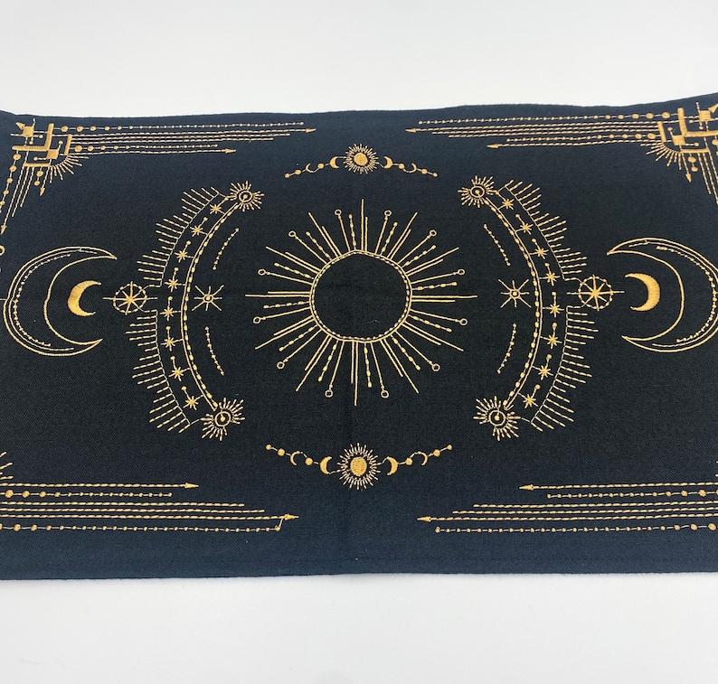 Puede incluir: Tela rectangular negra con un dise&ntilde;o celestial dorado. El dise&ntilde;o incluye un sol central, lunas crecientes, estrellas y formas geom&eacute;tricas. La tela probablemente se utiliza para el tarot u otras pr&aacute;cticas espirituales.