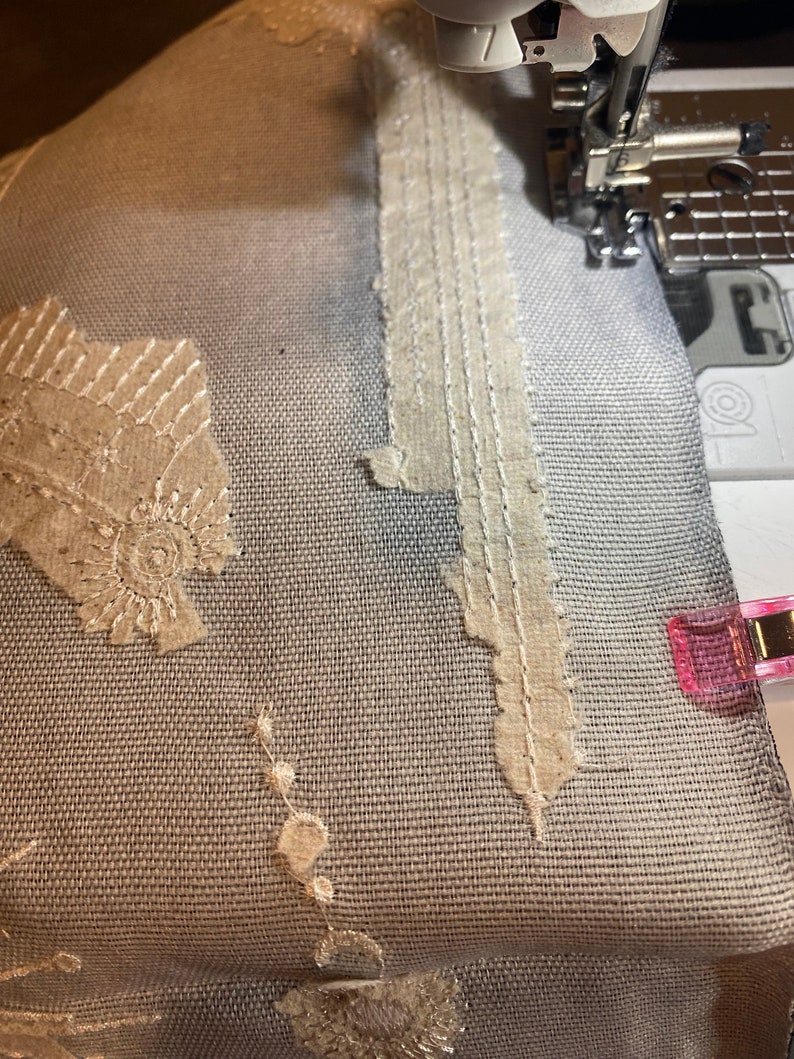 Puede incluir: Primer plano de un proyecto de costura en curso. Una tela azul claro est&aacute; adornada con encaje beige y costuras decorativas. Se ve una aguja de m&aacute;quina de coser, junto con una pinza rosa.