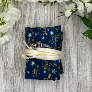May include: A folded, dark blue fabric item with a celestial pattern of stars and branches, secured with a cream-colored ribbon. A small crystal is attached to the ribbon. The background is a light wood surface with white flowers.