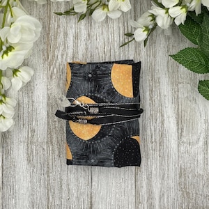 May include: A folded fabric item with a celestial pattern of black, gold, and gray moons and stars. A black ribbon with silver accents wraps around the item, secured with a clear crystal charm. The item is set against a light wood background.