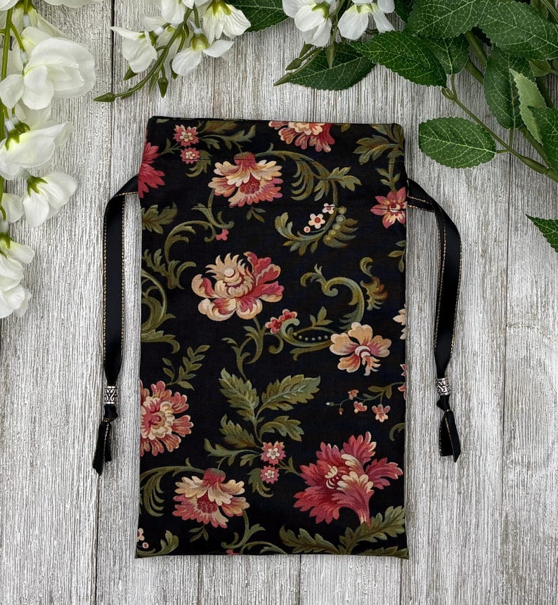 May include: A black drawstring bag with a floral pattern in shades of pink, cream, and green. The bag has black satin drawstrings with silver beads at the ends.