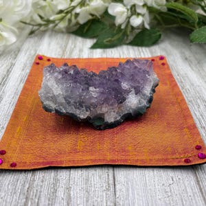 May include: A large amethyst crystal cluster resting on a small orange and red square cushion with pink rhinestones around the edges.