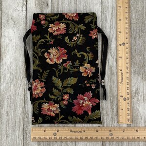 May include: A black fabric pouch with a floral print in shades of pink, green, and gold. The pouch has a drawstring closure and black ribbon ties.