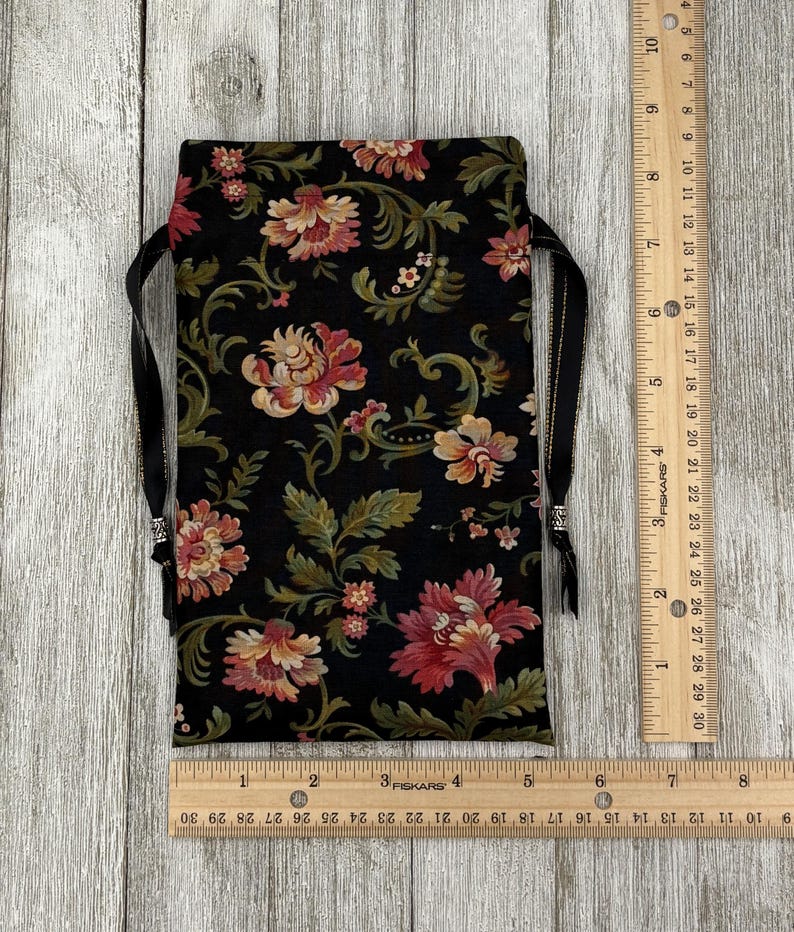 May include: A black fabric pouch with a floral print in shades of pink, green, and gold. The pouch has a drawstring closure and black ribbon ties.