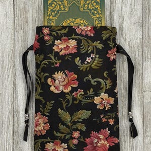 May include: A black drawstring bag with a floral pattern in shades of pink, green, and cream. The bag is made of fabric and has a black drawstring closure. The bag is filled with a green and gold deck of cards.