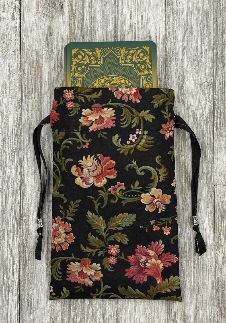 May include: A black drawstring bag with a floral pattern in shades of pink, green, and cream. The bag is made of fabric and has a black drawstring closure. The bag is filled with a green and gold deck of cards.