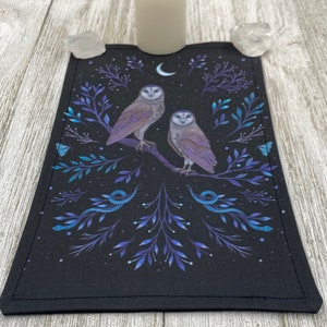 May include: Black rectangular altar cloth featuring two owls perched on a branch, with a crescent moon and botanical details in blue and purple. A white candle and two clear quartz crystals are on top.