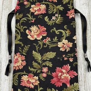 May include: Black fabric drawstring bag with a floral pattern in shades of pink, cream, and green. The bag has black ribbon ties with silver accents.