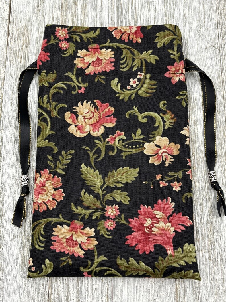 May include: Black fabric drawstring bag with a floral pattern in shades of pink, cream, and green. The bag has black ribbon ties with silver accents.