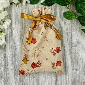 May include: A small, beige drawstring bag with a gold ribbon and a fall-themed print of pumpkins and leaves. The bag is tied with a gold ribbon and has silver beads on the drawstrings.