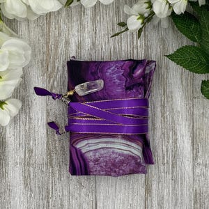 May include: A purple and white patterned fabric wrapped and tied with a purple ribbon. A clear crystal is attached to the ribbon. White flowers and green leaves are in the background.