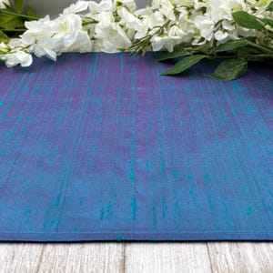 May include: A close-up of a blue and purple textile with a textured appearance. White flowers and green leaves are arranged at the top. The textile has a subtle sheen, suggesting a silk or similar material.