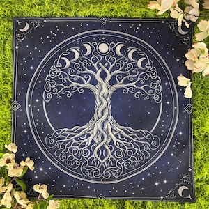 May include: A square, navy blue tapestry featuring a white, detailed illustration of a tree with intertwined roots and branches. The design is encircled by a ring of crescent moons and stars, set against a starry night sky. The tapestry is surrounded by a green surface and white flowers.
