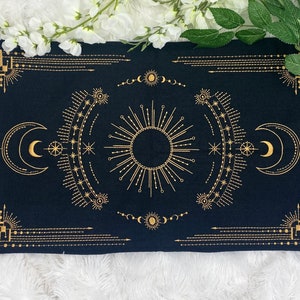 May include: Black rectangular tarot cloth with gold celestial design. The design includes a central sunburst, crescent moons, stars, and geometric borders. The cloth is likely used for tarot readings or spiritual practices.