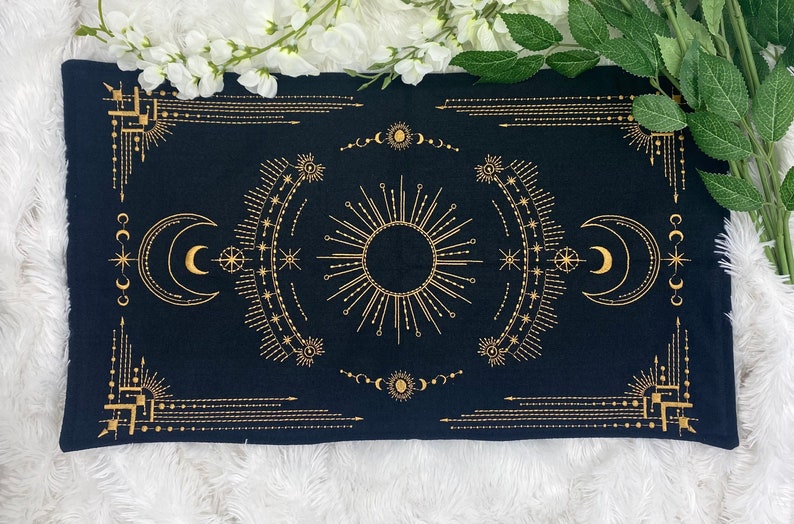 Puede incluir: Pa&ntilde;o de tarot rectangular negro con dise&ntilde;o celestial dorado. El dise&ntilde;o incluye un sol central, lunas crecientes, estrellas y bordes geom&eacute;tricos. El pa&ntilde;o se utiliza probablemente para lecturas de tarot.