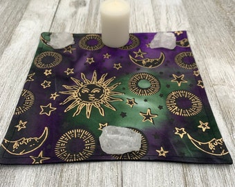 May include: A purple and green fabric altar cloth with a gold sun, moon, and stars design. There are three clear quartz crystals on the cloth and a white candle in the center.