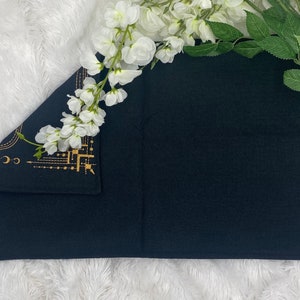 Puede incluir: Textil rectangular negro con dise&ntilde;o celestial dorado en la esquina. El textil est&aacute; doblado y colocado sobre una superficie blanca y esponjosa. Flores blancas y hojas verdes dispuestas alrededor del textil.