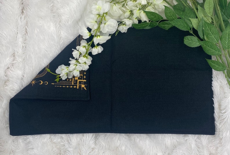 Puede incluir: Textil rectangular negro con dise&ntilde;o celestial dorado en la esquina. El textil est&aacute; doblado y colocado sobre una superficie blanca y esponjosa. Flores blancas y hojas verdes dispuestas alrededor del textil.