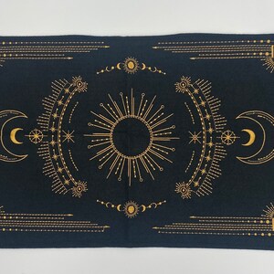 Puede incluir: Tapiz rectangular negro con un dise&ntilde;o celestial dorado. El dise&ntilde;o presenta un sol central, lunas crecientes, estrellas y patrones geom&eacute;tricos. El tapiz es probablemente de tela y es adecuado para la decoraci&oacute;n de paredes o como mantel.