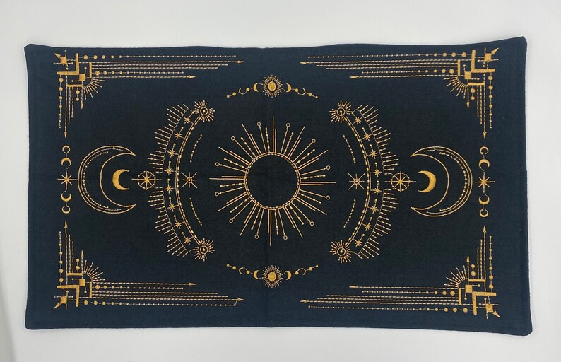 Puede incluir: Tapiz rectangular negro con un dise&ntilde;o celestial dorado. El dise&ntilde;o presenta un sol central, lunas crecientes, estrellas y patrones geom&eacute;tricos. El tapiz es probablemente de tela y es adecuado para la decoraci&oacute;n de paredes o como mantel.