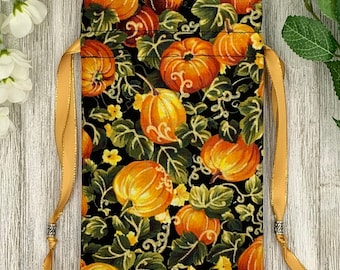 May include: A black fabric drawstring bag with a gold ribbon and a pumpkin pattern. The bag is filled with orange pumpkins and green leaves.
